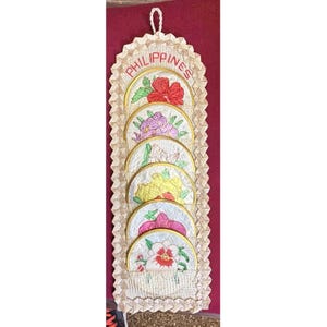 Può includere: Un'applique da parete decorativa con sei sottobicchieri floreali ricamati. I sottobicchieri sono disposti verticalmente in un supporto beige intrecciato. La parola "PHILIPPINES" è ricamata in rosso in alto.