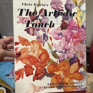 Puede incluir: Un libro de tapa dura titulado "The Artistic Touch" de Chris Unwin. La portada presenta un diseño floral en acuarela en tonos naranja, rosa, morado y amarillo. El subtítulo del libro dice "65 Outstanding Artists' Ideas & Creative Techniques".