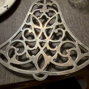 Pode incluir: Trivet de metal prateado em forma de sino com um intrincado design vazado. O trivet apresenta uma alça na parte superior e um padrão decorativo de linhas onduladas e formas florais. Este artigo de cozinha foi projetado para proteger as superfícies.