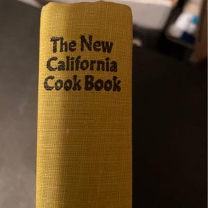 May include: A close-up of a cookbook spine. The book has a mustard yellow cover with the title "The New California Cook Book" printed in black. The cover has a textured appearance.
