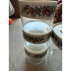 May include: A stack of three clear glass mixing bowls with a white rim featuring a colourful botanical pattern of various fruits and vegetables. Each bowl has a smooth, transparent body and a wide opening, designed for food preparation and storage.