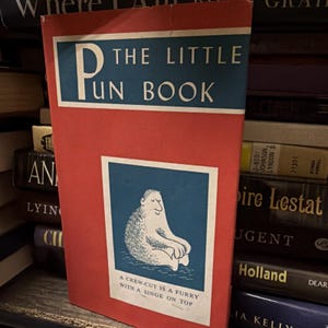 May include: A vintage book titled "The Little Pun Book" with a red cover and blue accents. The cover features a cartoon illustration of a furry creature. The text on the cover reads "The Little Pun Book".