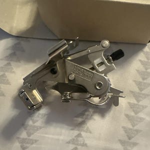 Könnte beinhalten: Ein silberfarbener Kräuselapparat für eine Nähmaschine, mit einem schwarzen Knopf und verschiedenen Hebeln und Metallkomponenten. Der Aufsatz befindet sich auf einer gemusterten Oberfläche, mit einem Pappkarton im Hintergrund. Auf der Box steht "RUFFLER HS LP".