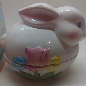 May include: A white ceramic bunny-shaped trinket box with pink-lined ears, blue eyes, and rosy cheeks. The box is decorated with colorful tulips in pink, blue, and yellow. A charming Easter-themed decorative item.