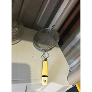 Pode incluir: Um coador de cozinha redondo com uma taça de malha metálica fina e um cabo com uma secção superior amarelo-pálido, uma faixa preta e uma base de cor creme com um orifício circular para pendurar. A estrutura metálica liga a malha ao cabo.