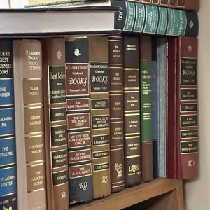 Könnte beinhalten: Eine Sammlung alter Bücher auf einem Holzregal, darunter Titel wie "Reader's Digest Condensed Books" und "Best Sellers". Die Buchrücken sind in verschiedenen Farben gehalten und weisen goldene Schriftzüge und dekorative Elemente auf.