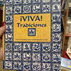 May include: A hardcover book titled ¡VIVA! Tradiciones, South Texas Cooks from Brush to Bay. The cover features a yellow central panel with blue text, surrounded by a grid of blue and white square tiles depicting various icons like animals.