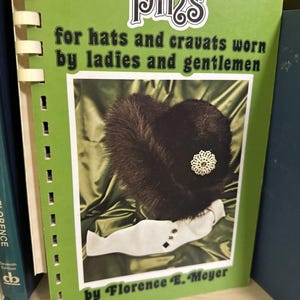 May include: A green book titled "Pins" with the subtitle "for hats and cravats worn by ladies and gentlemen." The cover features a dark fur hat with a decorative brooch. The author's name, Florence E. Meyer, is at the bottom.