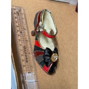 Peut inclure: Une chaussure à talons hauts miniature décorative. La chaussure présente un nœud noir avec un centre en strass et un design multicolore rouge, noir, vert et gris. La chaussure a une bordure dorée et un petit talon.