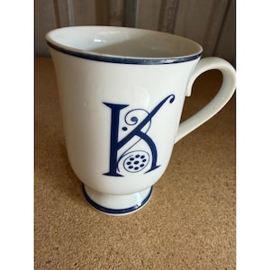Può includere: Una tazza in ceramica bianca con un bordo blu scuro e una grande lettera K stilizzata in blu scuro sulla parte anteriore. La tazza presenta una base a piedistallo con una fascia blu scuro e un manico curvo sul lato destro. La lettera K include volute.