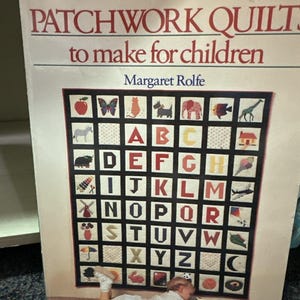 May include: A book titled "PATCHWORK QUILTS to make for children" by Margaret Rolfe. The cover features a colorful quilt design with the alphabet and various illustrations. A child is lying on the floor near the book.
