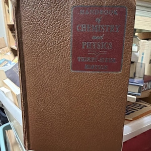 Puede incluir: Taza de cerámica blanca con asa negra y la frase "But First, Coffee" en negro. La taza se muestra sobre un fondo borroso, sugiriendo un ambiente acogedor.