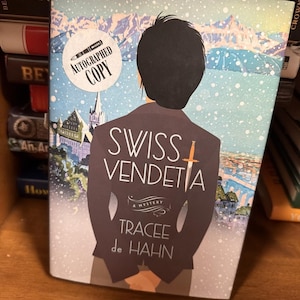 May include: A book titled "Swiss Vendetta" by Tracee de Hahn. The cover features a person's back with a sword and the title. The background shows a snowy landscape with buildings and mountains. An "Autographed Copy" sticker is on the cover.