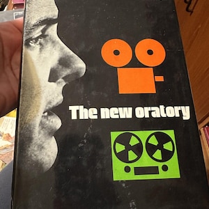 Könnte beinhalten: Ein schwarzer Buchumschlag mit dem Titel "The new oratory" in weißer Schrift. Der Umschlag zeigt ein Schwarzweißbild eines Männergesichts und zwei grafische Illustrationen einer Filmkamera und eines Tonbandgeräts.