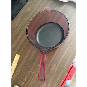 Puede incluir: Un cazo de cristal morado translúcido de forma redonda con un mango largo y un interior oscuro. Una regla de madera está colocada junto al cazo, mostrando una medida de aproximadamente 18 cm de diámetro para este recipiente de cocina.