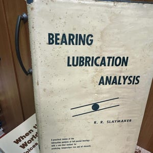 Puede incluir: Un libro antiguo titulado "BEARING LUBRICATION ANALYSIS" con un asa negra. La portada es de color blanquecino con texto negro y un diagrama. El nombre del autor es R. R. Slaymaker. El libro está en una estantería con otros libros.