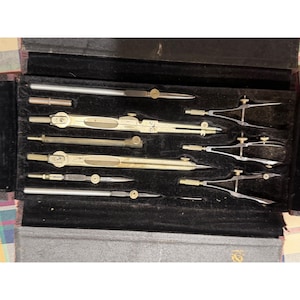 Puede incluir: Un juego de instrumentos de dibujo técnico vintage presentado en un estuche forrado de terciopelo negro. La colección incluye varios compases, divisores y barras de extensión de metal en tonos latón y plata con tornillos ajustables.