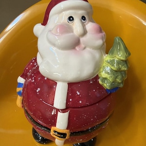 May include: A ceramic Santa Claus figurine with a red suit, white beard, and a green Christmas tree. The Santa Claus has a white hat with a red trim and a black belt with a gold buckle. The figurine is on a yellow surface.