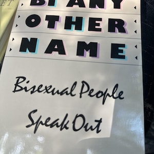 Op de afbeelding: Boekomslag met de titel "BI ANY OTHER NAME" in vette zwarte letters met een lichtblauw en paars schaduweffect. De ondertitel "Bisexual People Speak Out" is in handschrift.