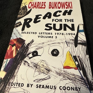 May include: Book cover for "Reach for the Sun: Selected Letters 1978-1994, Volume 3" by Charles Bukowski. The cover features a black and white illustration of a face with a yellow and red background.