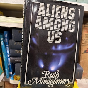 May include: A hardcover book titled "Aliens Among Us" by Ruth Montgomery. The book cover is black with white text and a blue and white abstract design. The book is displayed on a shelf with other books.