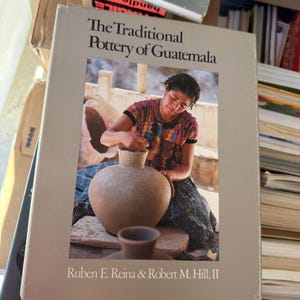 Puede incluir: Portada del libro "The Traditional Pottery of Guatemala" con una foto de una mujer trabajando en una olla de barro. La portada es de color beige claro con texto negro. Los nombres de los autores están en la parte inferior.