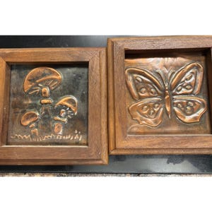 May include: Two square wall plaques featuring embossed copper metal art set within dark brown wooden frames. One plaque displays a cluster of three mushrooms, while the other plaque depicts a detailed butterfly with textured wing patterns.