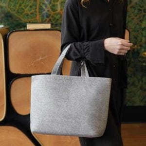 May include: A light gray tote bag with two handles. The bag is made of a felt-like material and has a rectangular shape. The bag is being held by a person wearing a black dress.