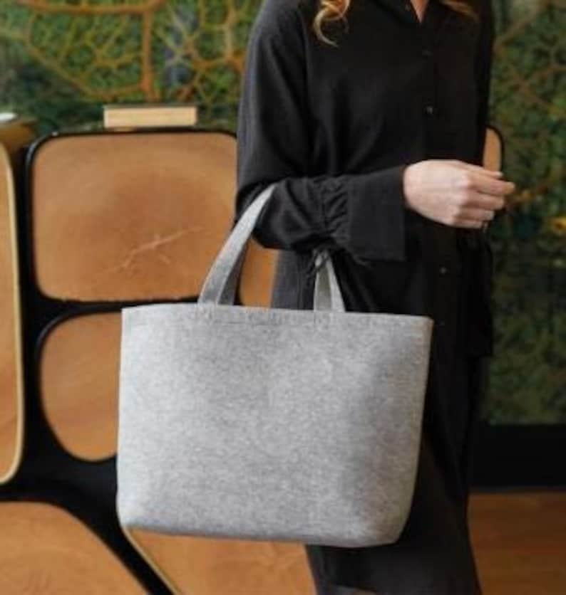 May include: A light gray tote bag with two handles. The bag is made of a felt-like material and has a rectangular shape. The bag is being held by a person wearing a black dress.