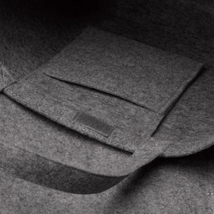May include: A close-up of a gray felt tote bag with a rectangular pocket and a handle. The bag is made of a soft, textured material. The pocket has a small, rectangular patch sewn on it.