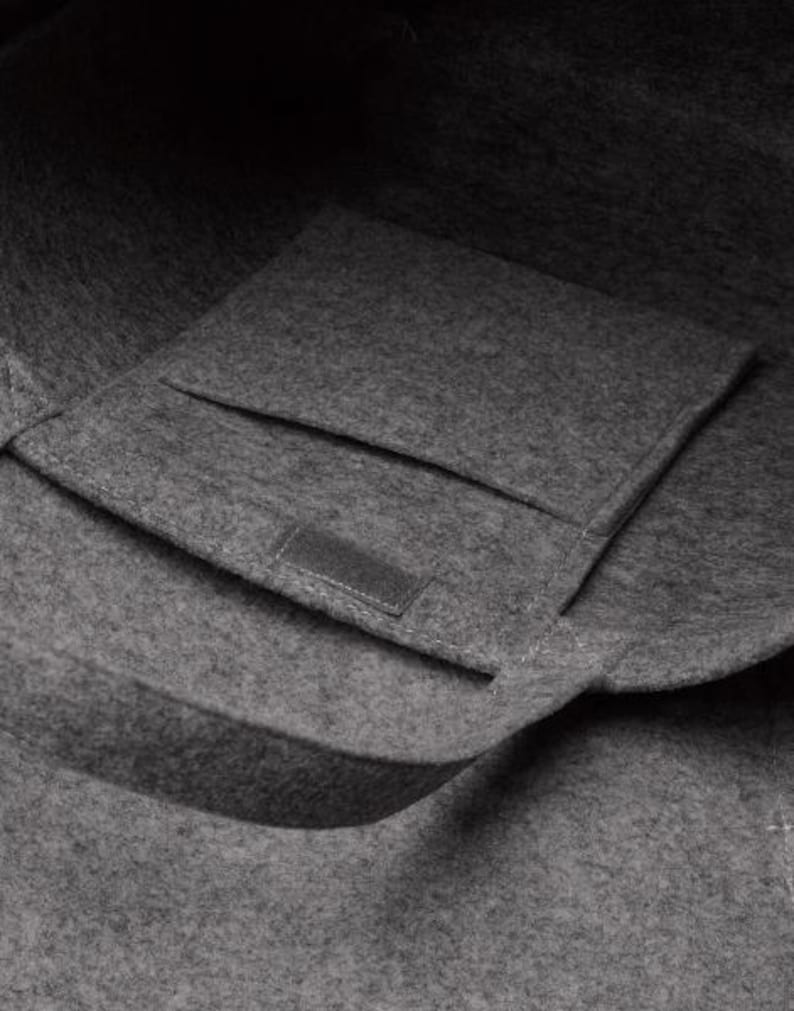 May include: A close-up of a gray felt tote bag with a rectangular pocket and a handle. The bag is made of a soft, textured material. The pocket has a small, rectangular patch sewn on it.