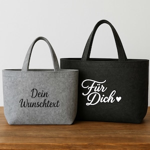 Könnte beinhalten: Zwei Tragetaschen, eine graue und eine schwarze, mit Henkeln. Die graue Tasche hat den Text "Dein Wunschtext" in schwarzer Schrift. Die schwarze Tasche hat den Text "Für Dich" mit einem Herzsymbol in weißer Schrift. Beide Taschen sind aus Filz.