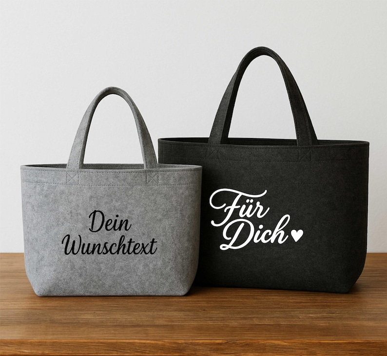 May include: Two tote bags, one gray and one black, with handles. The gray bag has the text "Dein Wunschtext" in black script. The black bag has the text "F&uuml;r Dich" with a heart symbol in white script. Both bags are made of a felt-like material.