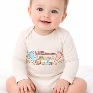 May include: A baby wearing a long-sleeved, off-white onesie. The onesie has the text "Willkommen kleines Wunder" in pink and yellow, with blue and pink footprints and star and heart accents.