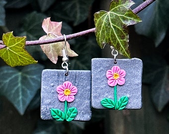 May include: A pair of gray square earrings with pink flowers and green leaves. The earrings are hanging from a branch.