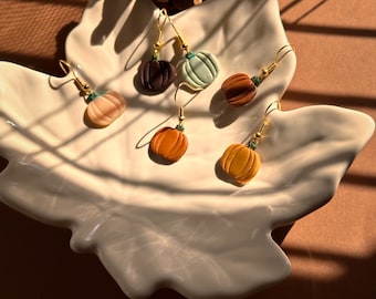 Pode incluir: Cinco pares de brincos em forma de abóbora num prato branco. Os brincos são feitos de argila polimérica e têm cores diferentes, incluindo castanho, verde, laranja e pêssego.