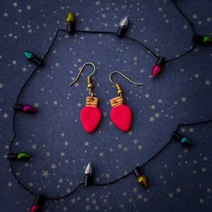 Puede incluir: Un par de pendientes de bombilla rosa con detalles dorados. Los pendientes cuelgan de una cadena de luces negras con bombillas de colores.