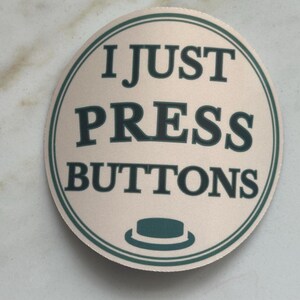 Może przedstawiać: Okrągła, beżowa naklejka z napisem "I JUST PRESS BUTTONS" w ciemnozielonym kolorze. Naklejka ma ciemnozieloną obw&oacute;dkę i małą grafikę kapelusza na dole. Naklejka znajduje się na jasnej, marmurowej powierzchni.