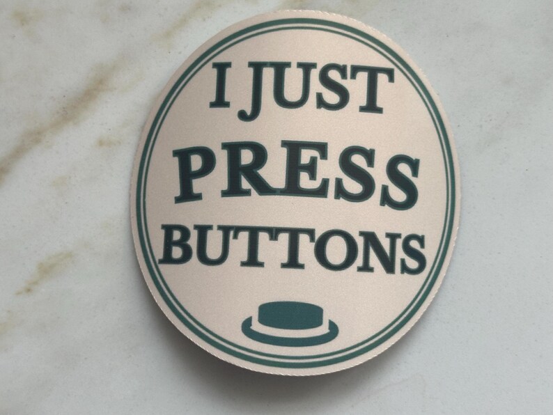 Może przedstawiać: Okrągła, beżowa naklejka z napisem "I JUST PRESS BUTTONS" w ciemnozielonym kolorze. Naklejka ma ciemnozieloną obw&oacute;dkę i małą grafikę kapelusza na dole. Naklejka znajduje się na jasnej, marmurowej powierzchni.