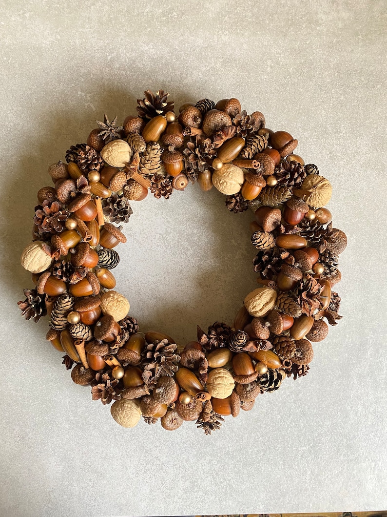May include: A brown, circular wreath made of pinecones, acorns, walnuts, and cinnamon sticks. The wreath is decorated with gold beads.