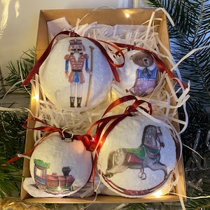 May include: Four white Christmas baubles with red ribbon. The baubles feature a nutcracker, a teddy bear, a rocking horse, and a toy train.