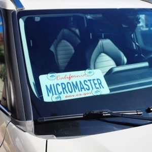 May include: A blue and white California license plate with the text "MICROMASTER" and "dmv.ca.gov" in red. The plate is attached to the windshield of a car.