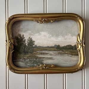 May include: A vintage landscape painting in a gold ornate frame. The artwork depicts a serene scene with a river, trees, and a cloudy sky. The frame has a rectangular shape with curved edges and decorative detailing, adding a touch of elegance.