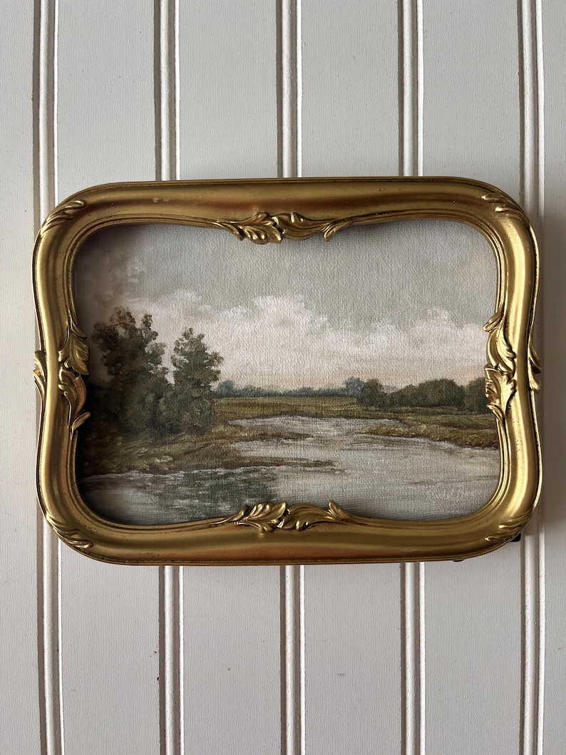 May include: A vintage landscape painting in a gold ornate frame. The artwork depicts a serene scene with a river, trees, and a cloudy sky. The frame has a rectangular shape with curved edges and decorative detailing, adding a touch of elegance.