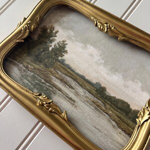 May include: A vintage-style framed landscape painting. The artwork depicts a river scene with trees and a cloudy sky. The ornate gold frame has a curved design with decorative leaf accents, adding a touch of elegance to the artwork.