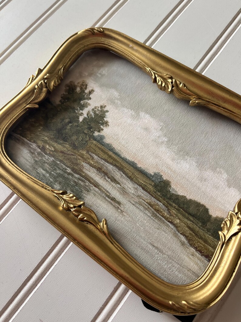 May include: A vintage-style framed landscape painting. The artwork depicts a river scene with trees and a cloudy sky. The ornate gold frame has a curved design with decorative leaf accents, adding a touch of elegance to the artwork.