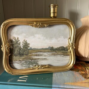 May include: A framed landscape painting with a gold ornate frame. The artwork depicts a serene waterside scene with trees and a cloudy sky. The frame is rectangular with decorative flourishes. The painting rests on a book titled "THE IMPRESSIONISTS".