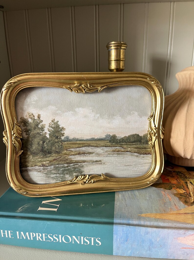 May include: A framed landscape painting with a gold ornate frame. The artwork depicts a serene waterside scene with trees and a cloudy sky. The frame is rectangular with decorative flourishes. The painting rests on a book titled "THE IMPRESSIONISTS".