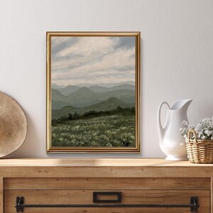 Peut inclure: Une peinture de paysage encadr&eacute;e dans un cadre dor&eacute; repr&eacute;sente des collines verdoyantes sous un ciel nuageux. Le premier plan montre un champ de fleurs blanches. Un chapeau de paille, un pichet blanc et un panier en osier avec des fleurs sont sur une surface en bois.