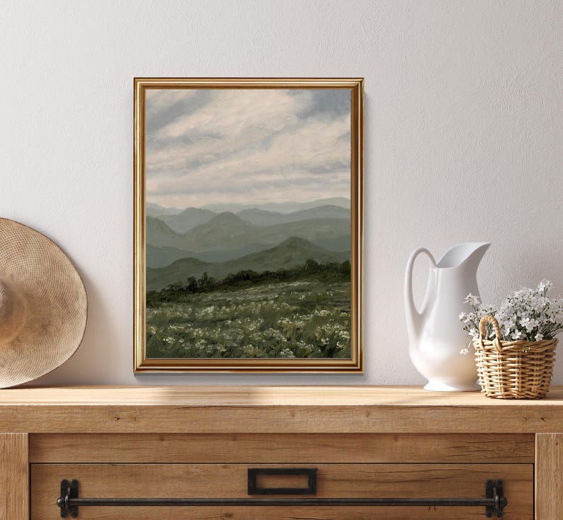 Peut inclure: Une peinture de paysage encadr&eacute;e dans un cadre dor&eacute; repr&eacute;sente des collines verdoyantes sous un ciel nuageux. Le premier plan montre un champ de fleurs blanches. Un chapeau de paille, un pichet blanc et un panier en osier avec des fleurs sont sur une surface en bois.